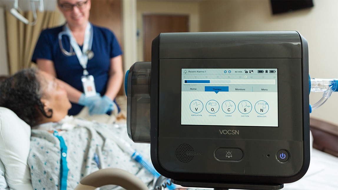 Nurse and patient in a hospital room with a ventilator in the foreground.
