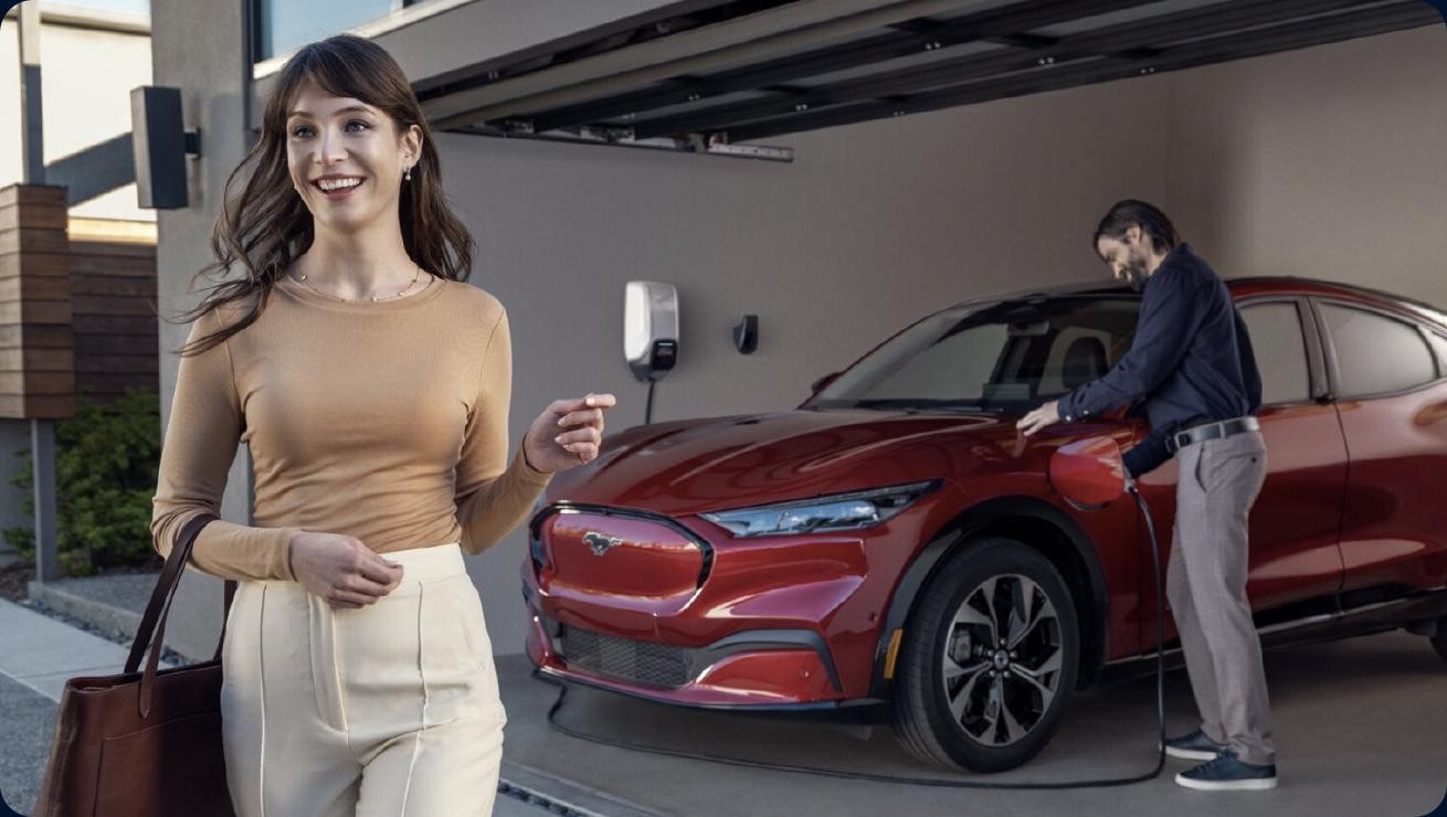 A woman walks away from a Mustang Mach-E as a man plugs it into a home charging station