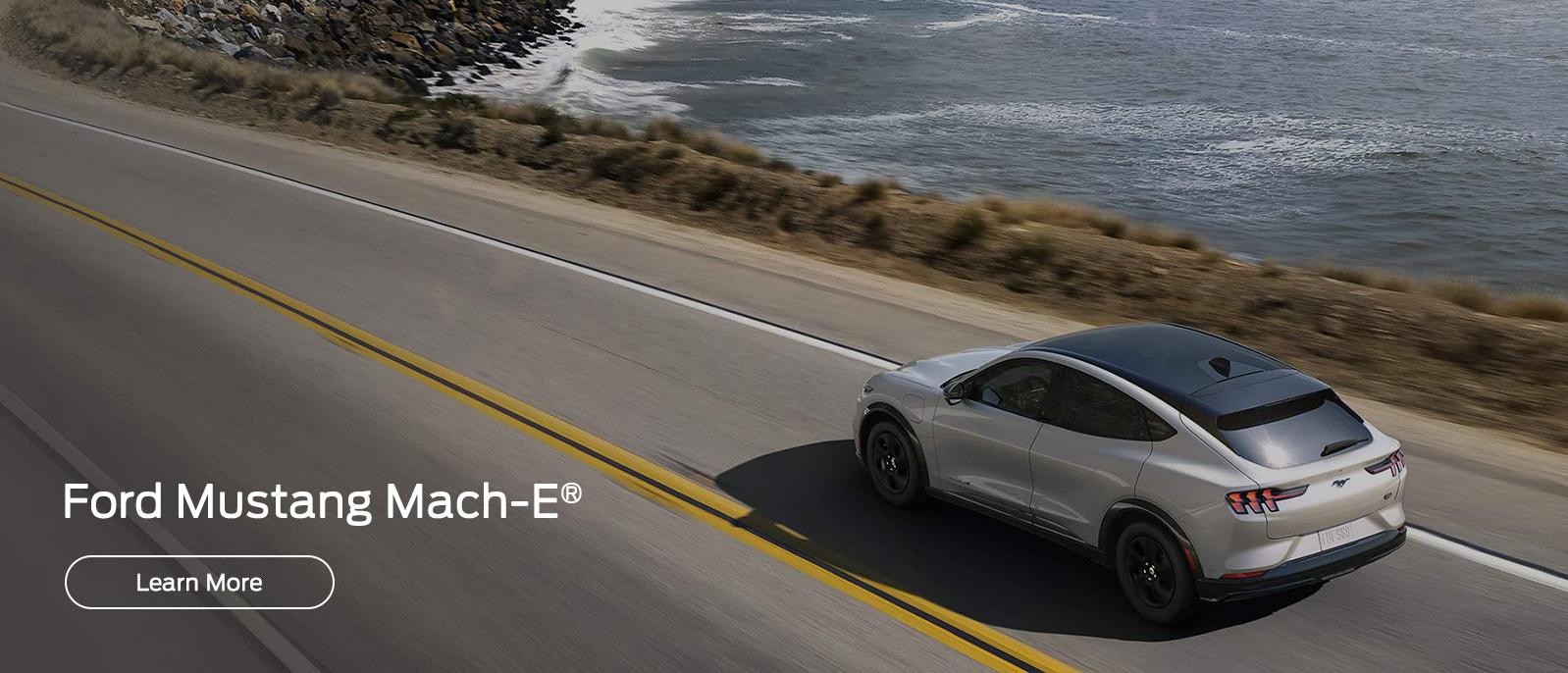 A Silver 2023 Ford Mustang Mach-E driving down the road along the ocean.