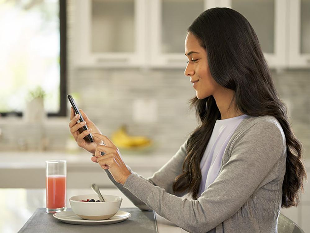 woman eating bowl of fruit with juice scrolling on her phone at the table