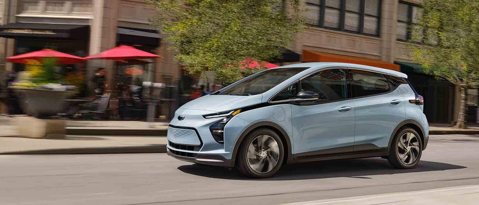 A light blue Chevy Bolt EV driving down a city street. A light blue Chevy Bolt EV driving down a city street.