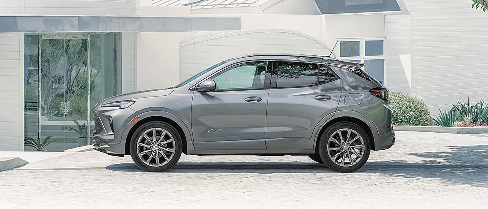 A 2024 Buick Encore GX Avenir in Moonstone Gray Metallic parked in the driveway of a crisp, white, modern house.