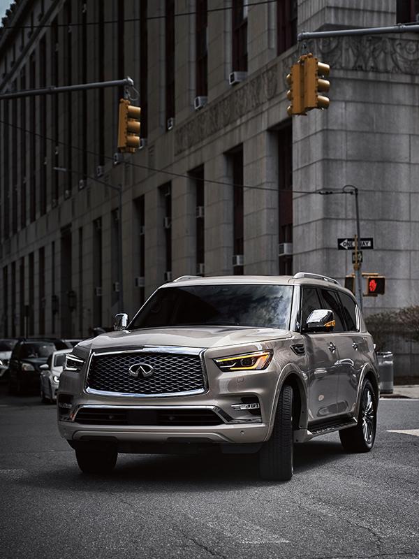 A tan INFINITI QX80 driving around a city street corner. A tan INFINITI QX80 driving around a city street corner.