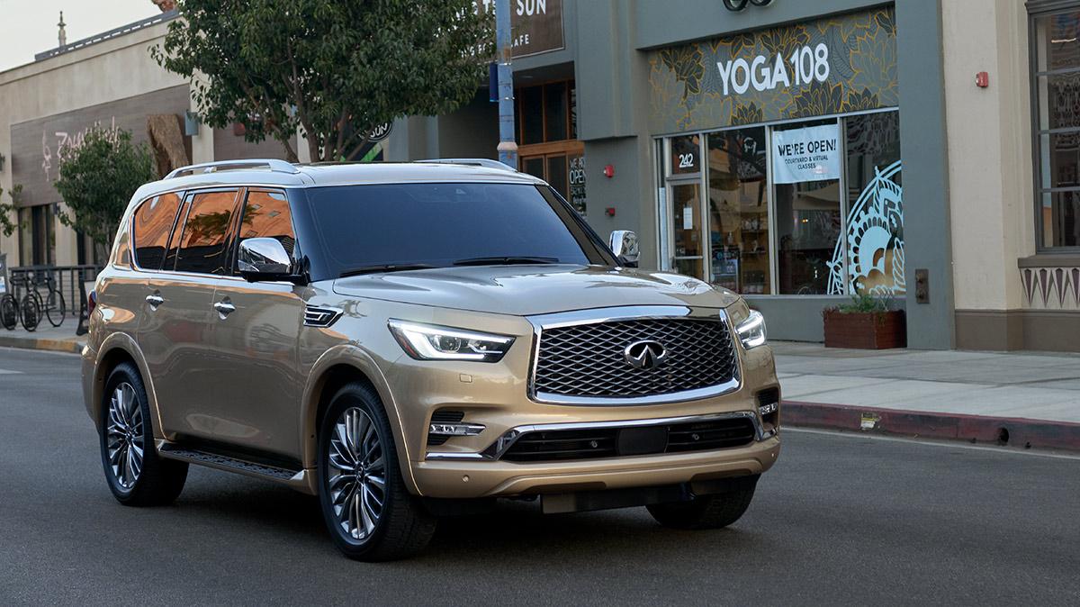 A tan INFINITI QX80 driving down a city street at sunset. A tan INFINITI QX80 driving down a city street at sunset.