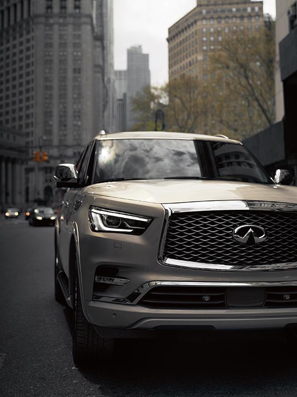 Nearly head-on view of a tan INFINITI QX80 driving through a city street. Nearly head-on view of a tan INFINITI QX80 driving through a city street.