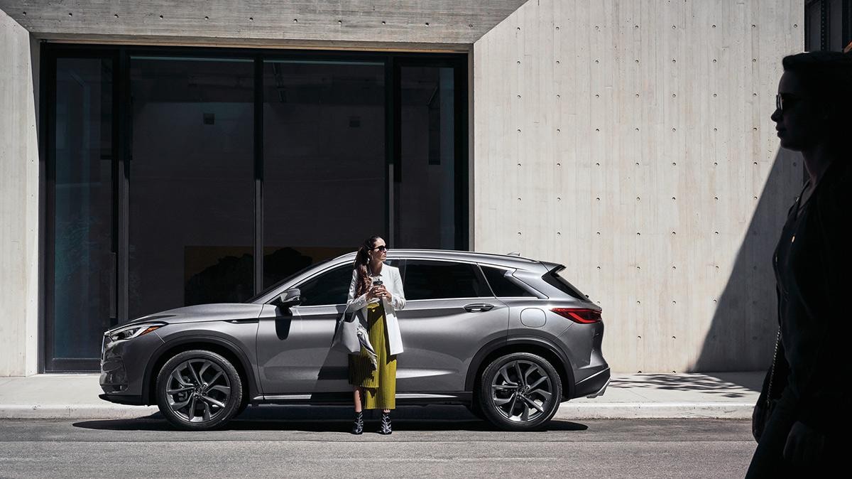 A woman standing in front of her grey INFINITI QX50 in front of an office building.