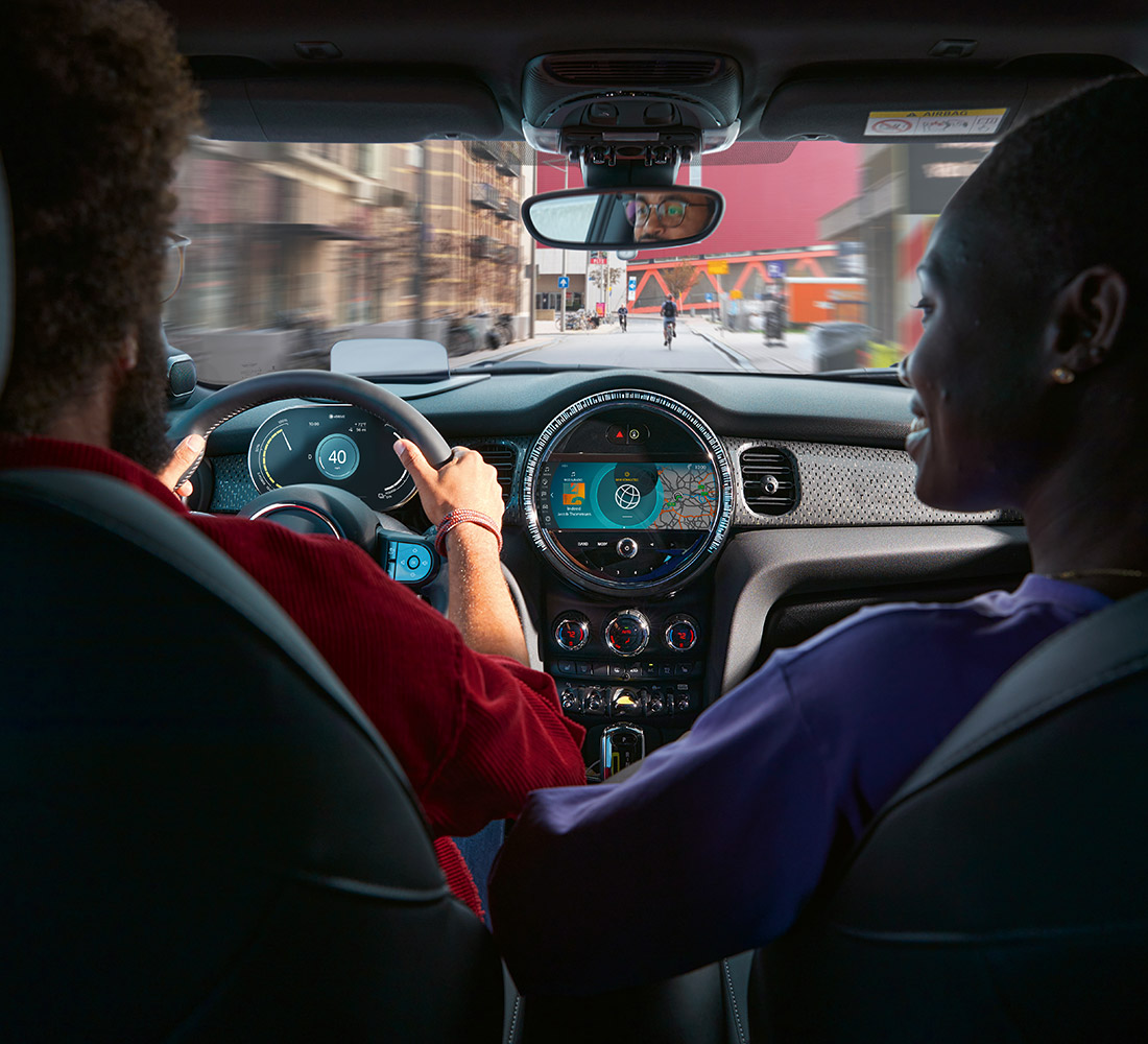A look over the right shoulder of a couple driving their 2022 MINI Cooper SE Hardtop 2 Door around the city.