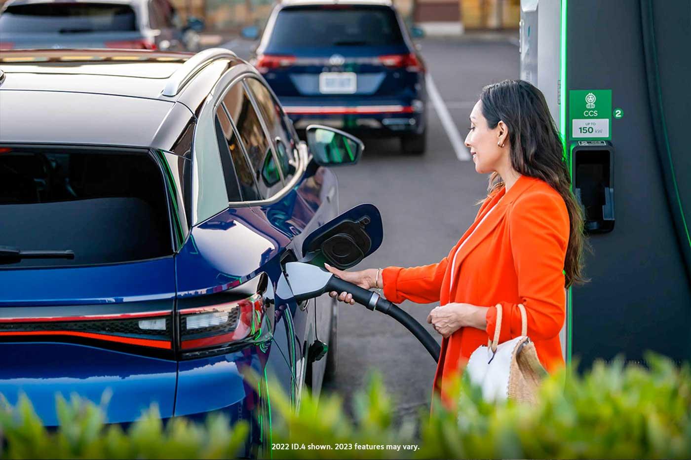 A well-dressed woman, parked at charging station stands on the passenger side of an ID.4 in Dusk Blue Metallic as she plugs in the car.
