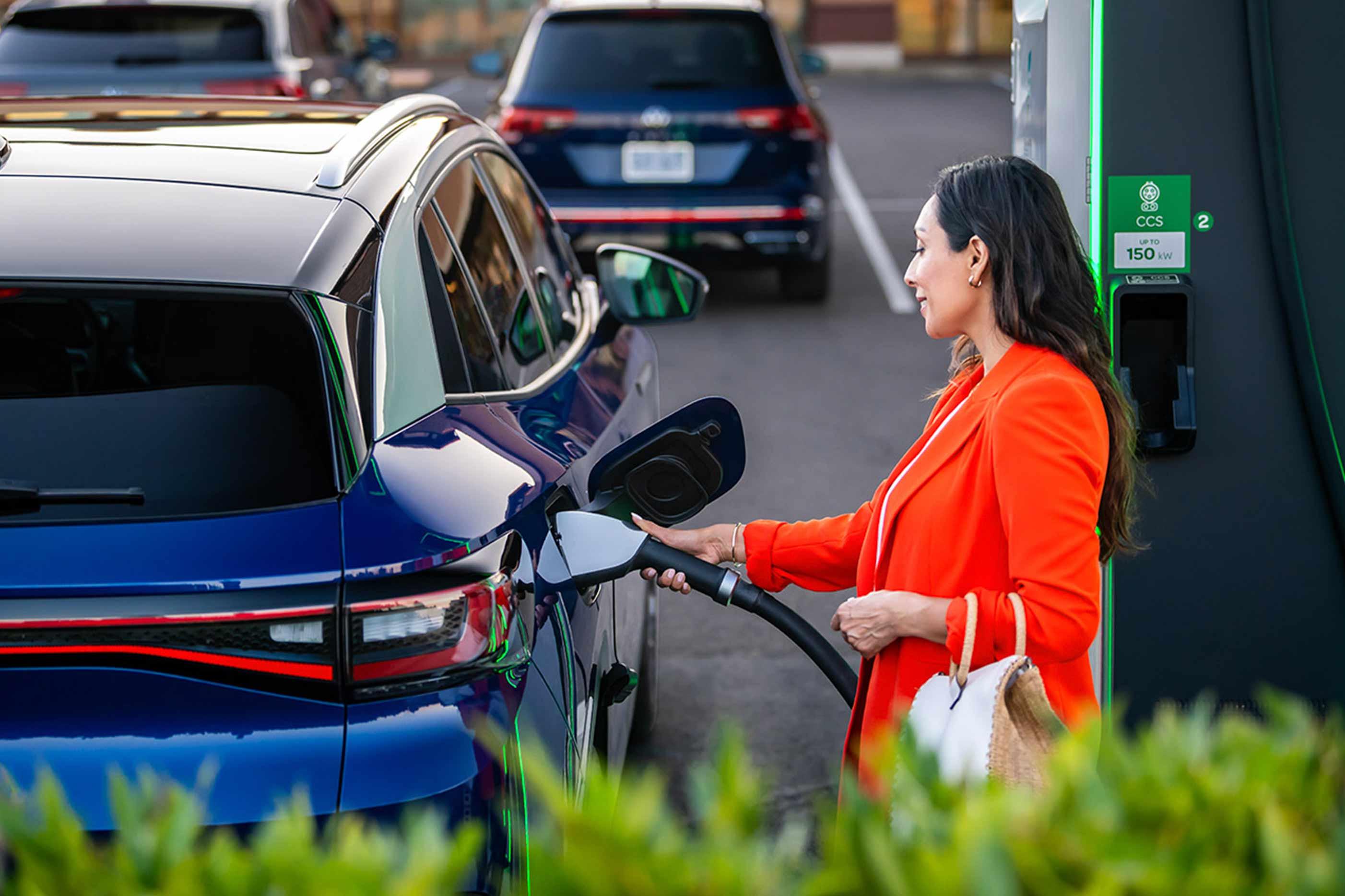A well-dressed woman, parked at charging station stands on the passenger side of an ID.4 in Dusk Blue Metallic as she plugs in the car.
