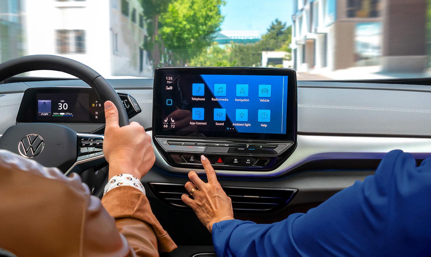 A close-up shot of an ID.4 SUV dashboard over the shoulders of the driver and passenger, focusing on the center display as thepassenger reaches out to the touchscreen.