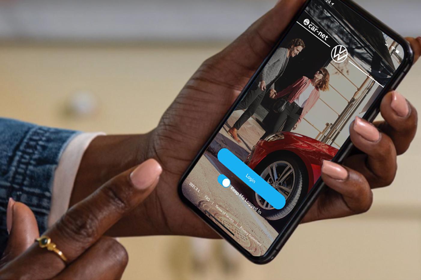 A hand holds a smartphone as a finger reaches to tap the VW Car-Net Login button on the screen