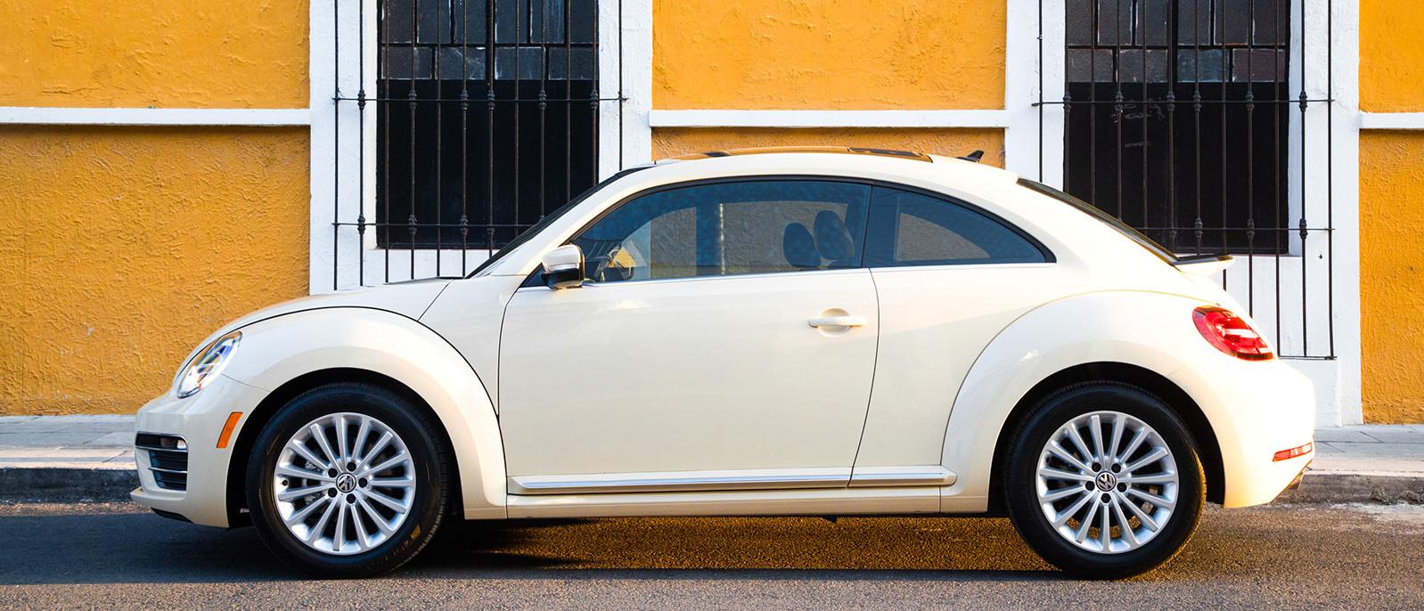 A cream-colored Volkswagen Beetle parked in front of a marigold orange wall with white-trimmed windows. A cream-colored Volkswagen Beetle parked in front of a marigold orange wall with white-trimmed windows.